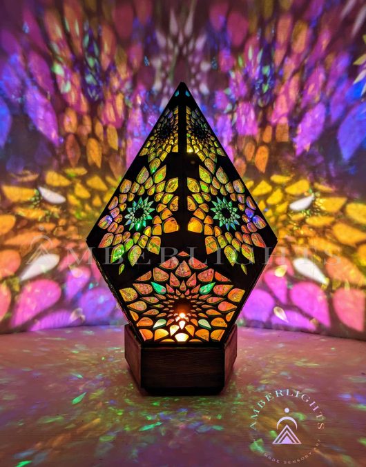 Handcrafted pyramid-shaped sensory lamp by AmberLights, adorned with intricate multicolored patterns casting vibrant kaleidoscopic projections onto surrounding surfaces.