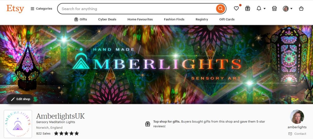 This screenshot showcases the AmberlightsUK Etsy shop, a top-rated store specializing in handmade sensory meditation lights and unique illuminated art pieces. The vibrant banner displays colorful, glowing designs, reflecting the shop's focus on creative and calming decor. Located in Norwich, England, the shop proudly highlights its 922 sales and a perfect 5-star review rating, building trust and credibility with potential customers. Recognized as a "Top shop for gifts," AmberlightsUK offers unique, high-quality products perfect for gifting or adding a warm, artistic touch to any space.